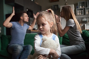 a little girl holding her tedy bear while her parents are arguing in the background