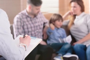 professional family psychotherapist writing notes in front of a couple with a child in a blurred background during a consultation