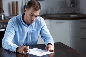 sad man in shirt sitting at table with ring and signing divorce documents 